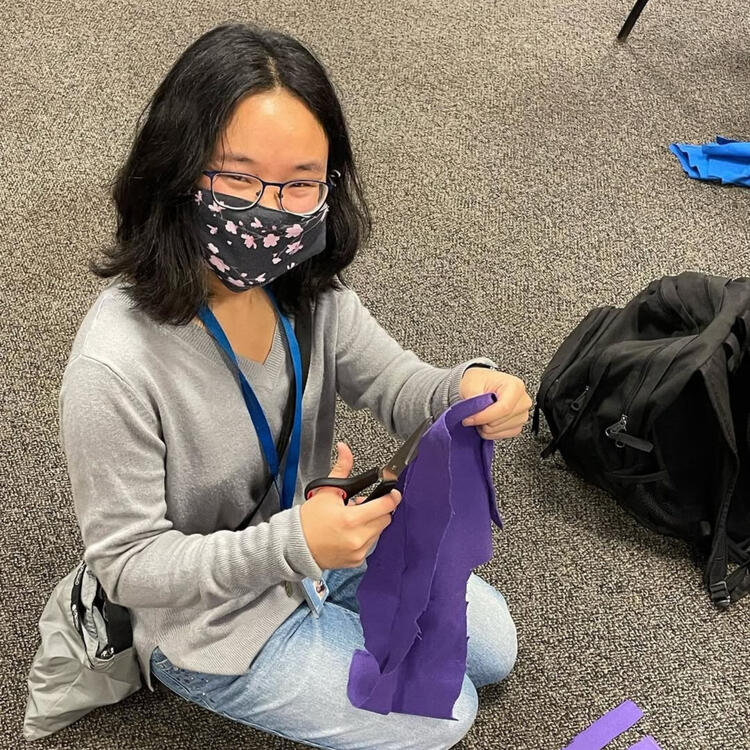 A person sits on the floor wearing a black mask with flowers and holding purple cloth that they are using scissors to cut.